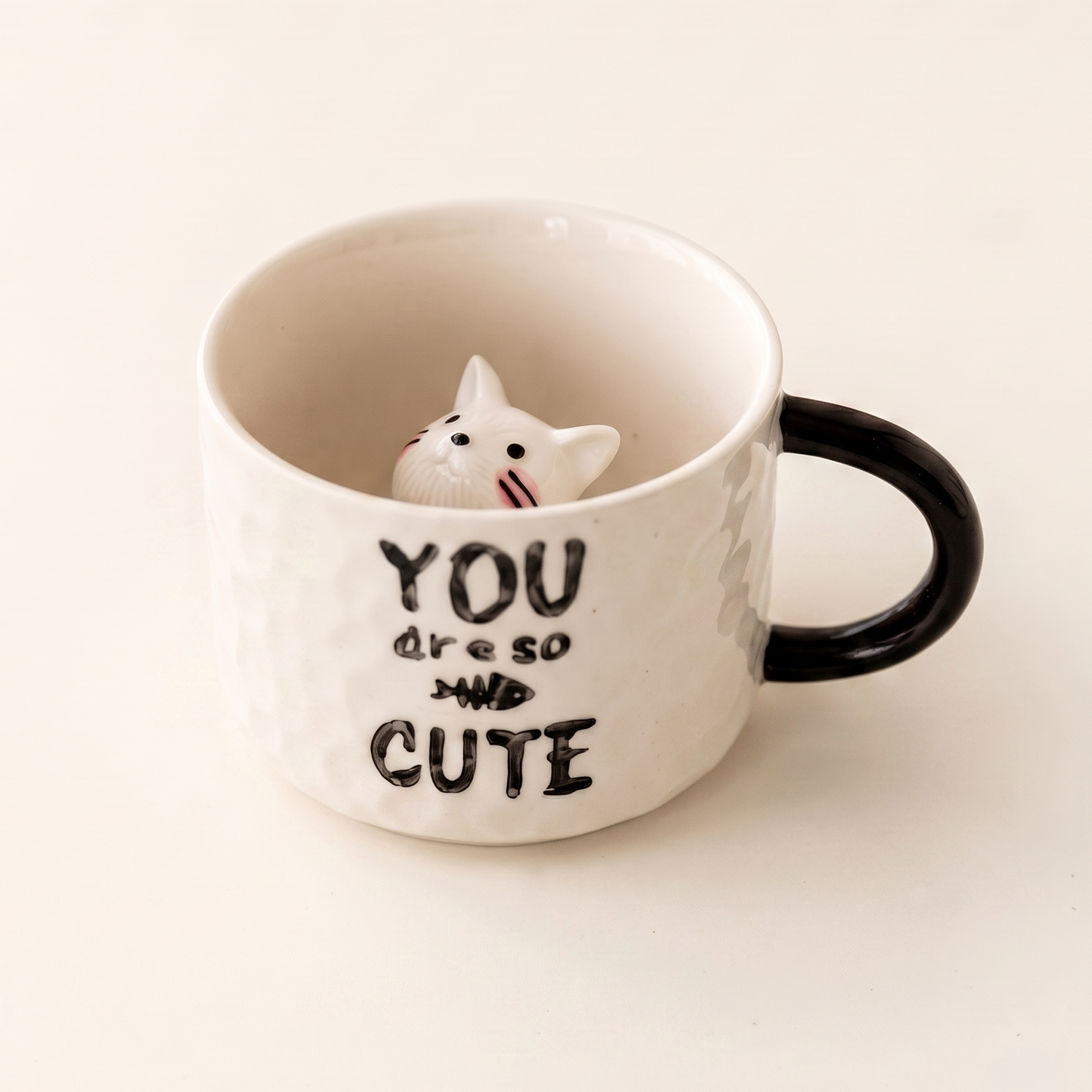 A white ceramic mug with a black handle and black text reading 'YOU are so CUTE' alongside a small white cat figurine peeking out of the inside of the mug, set against a plain off-white background.