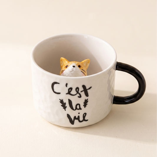 A white ceramic mug with a black handle and black printed French text reading 'C'est la vie' on a warm cream background. A small orange-and-white cat figurine is peeking out from inside the mug.